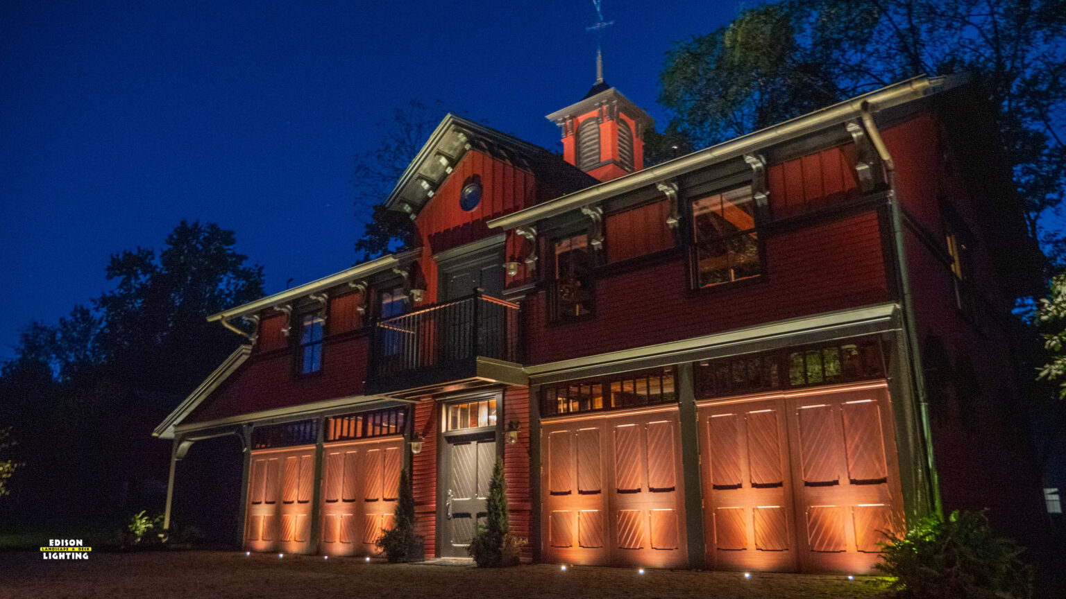 A beautifully illuminated carriage house featuring warm, low-voltage landscape lighting that highlights the architectural details and enhances curb appeal. The strategic placement of uplights along the garage doors creates depth and elegance, while accent lights on the upper-level facade add a refined touch.